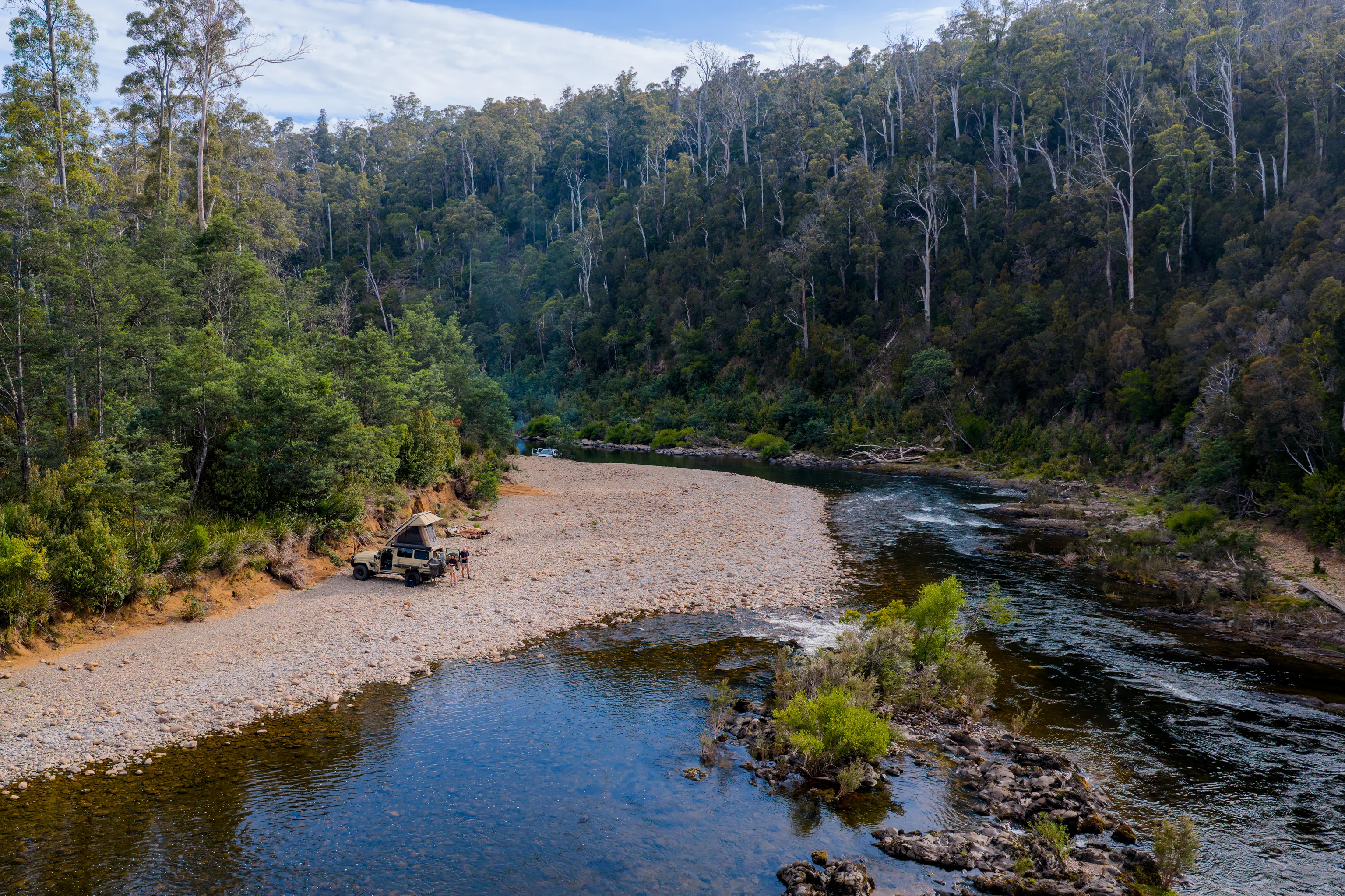 Aerial view of a 4WD with rooftop tent camped on a gravel riverbank in a forested gorge